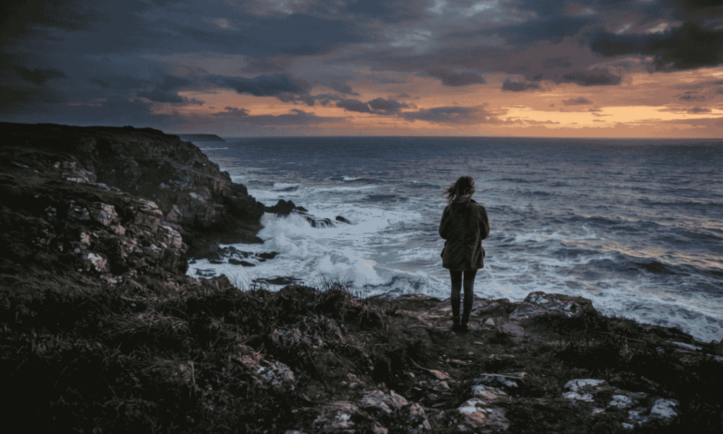 "A lonely figure standing on a grassy cliff overlooking a turbulent, dark ocean at sunset, with a dramatic, cloudy sky above."